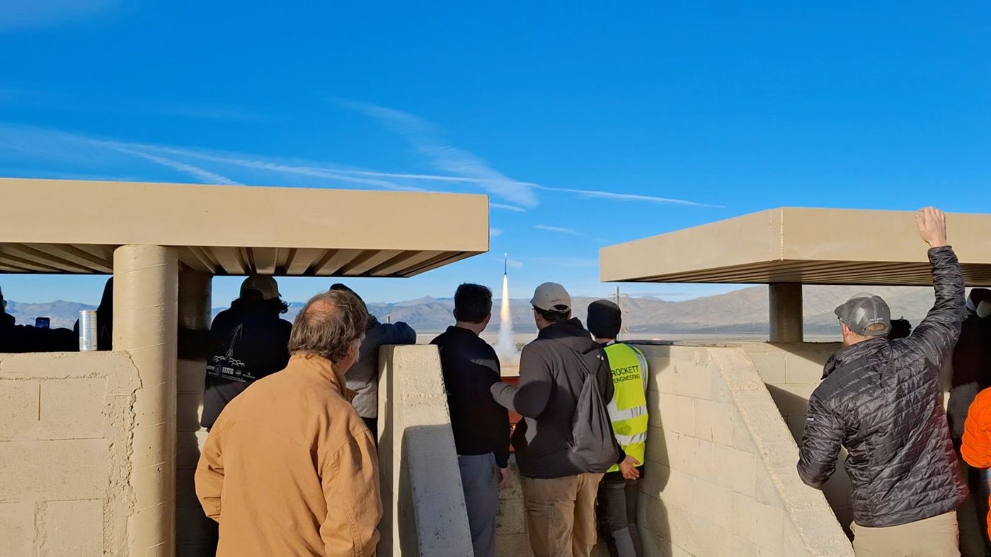 A launch vehicle lifting off from a desert launch pad, spectators watching from a concrete blockhouse in the foreground.
