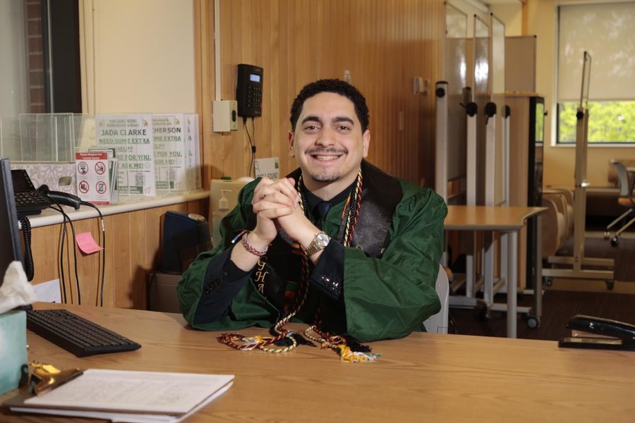 Kevin Polanco at the tutorial center front desk in graduation regalia, hands clasped, smiling.