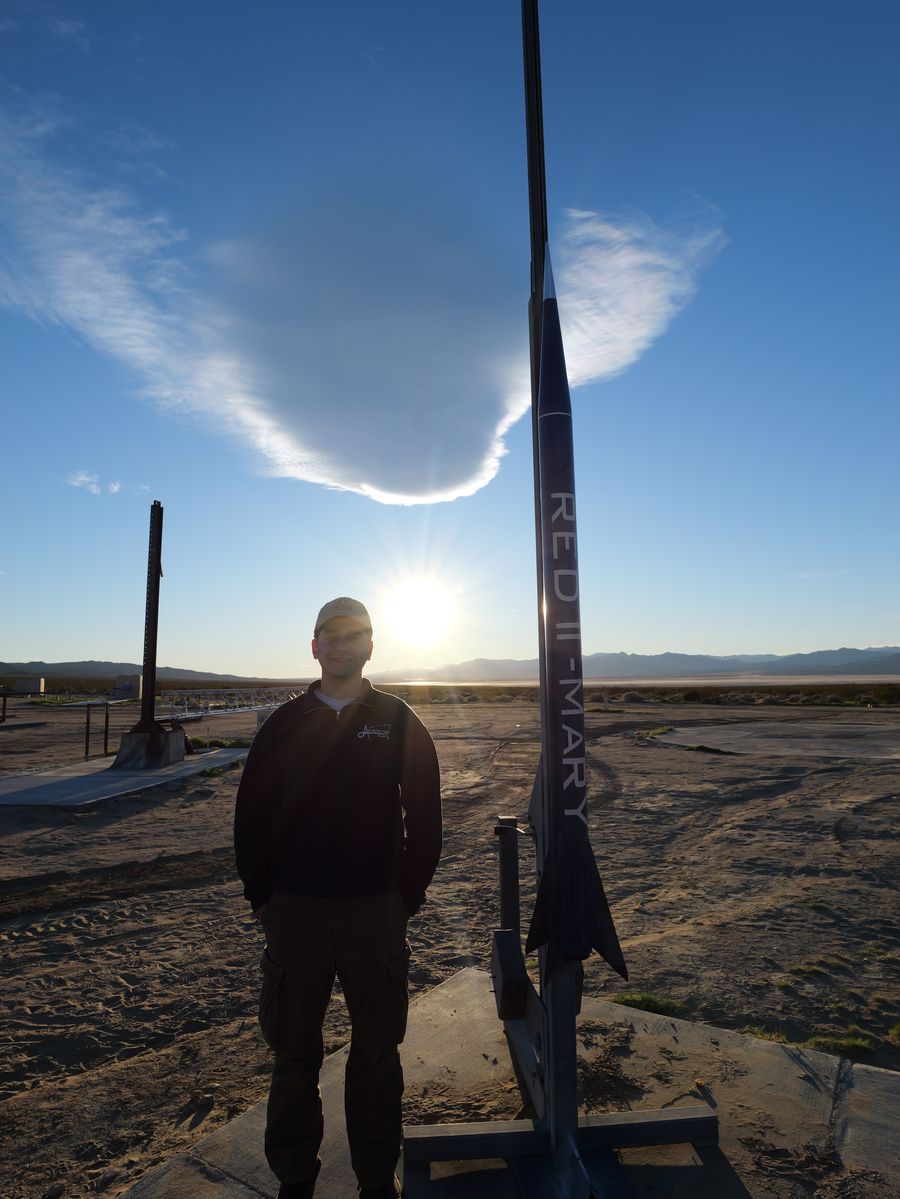 Kevin Polanco standing beside RED II Mary on the launch rail at a desert launch site, the sun low behind them.